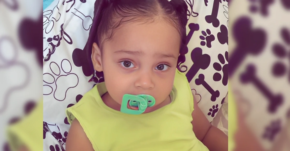 A young child with a green pacifier in the mouth, wearing a lime-green sleeveless shirt, resting on a blanket with paw and bone patterns in the background.