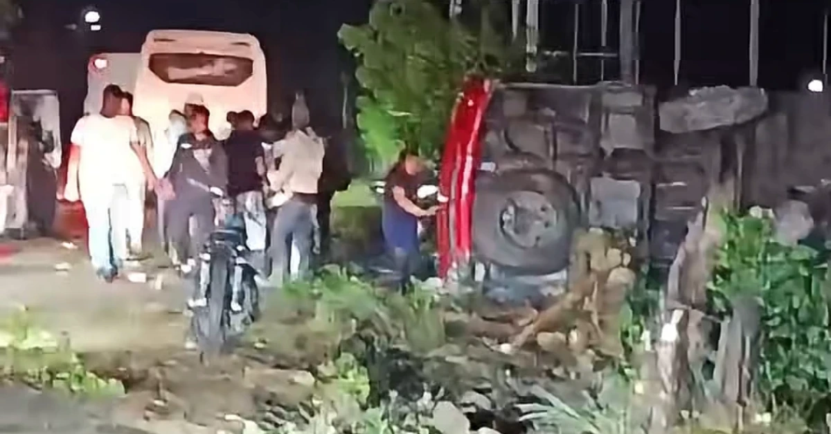 Nighttime scene with a red overturned vehicle and a group of people inspecting it near a concrete wall. Debris on the ground creates a rough roadside scene.