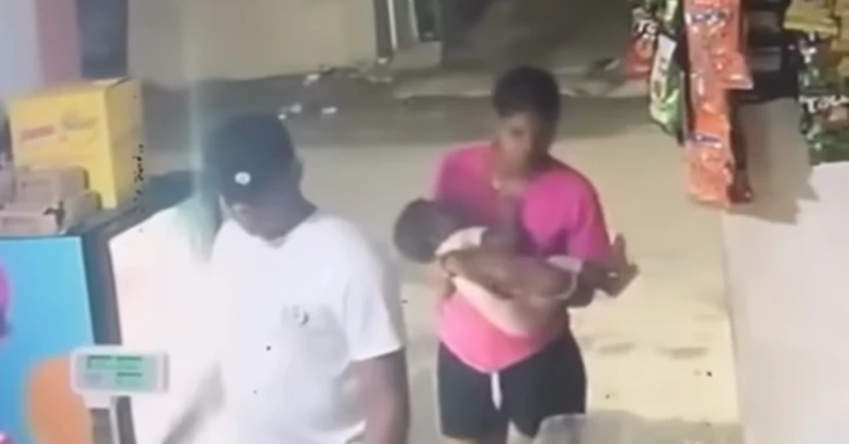 Man in white shirt and dark cap walks past a storefront as a woman in pink cradles a baby nearby.