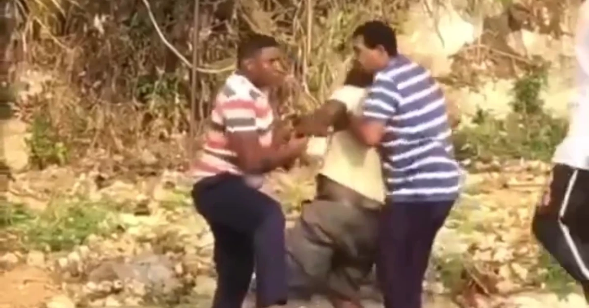 Two men restrain a kneeling man outdoors while another man watches nearby in a dusty, leafy area.