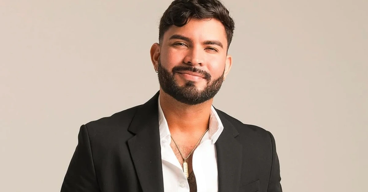 Smiling man with a beard in a black suit jacket and white shirt, wearing a pendant necklace, against a beige background.