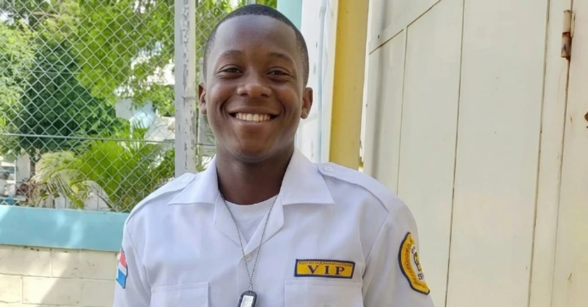 Smiling security guard in a white uniform with a VIP badge, standing outdoors beside a fence.