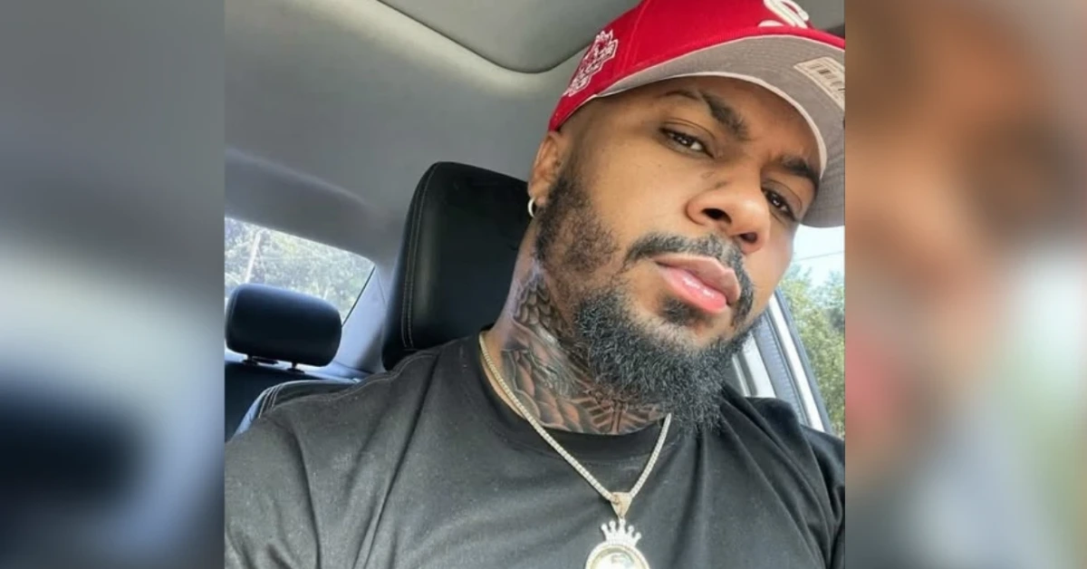 Close-up selfie of a man with a beard and neck tattoos wearing a red cap and gold chain in a car momentarily looking at the camera.