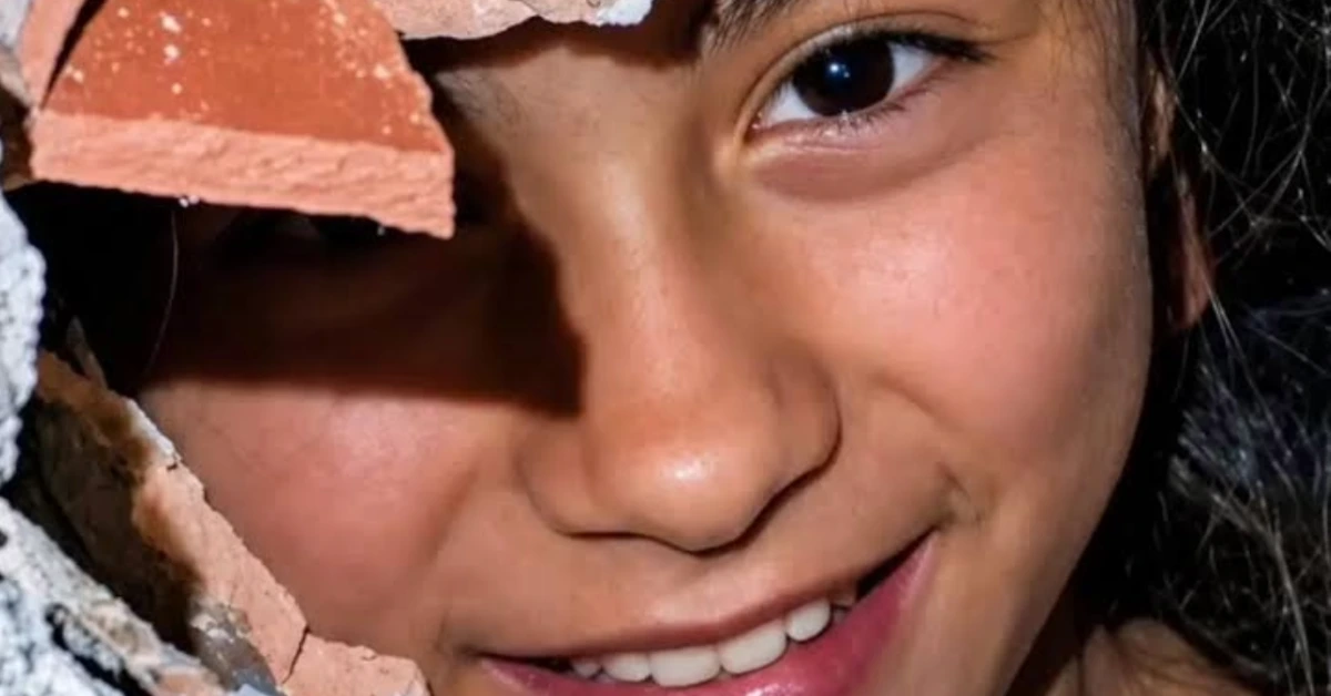 Close-up of a smiling girl’s face peeking through a crack in a brick wall.