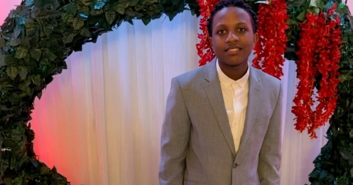 Young man in a light gray suit and white shirt posing in front of a floral arch with red hanging flowers behind him.