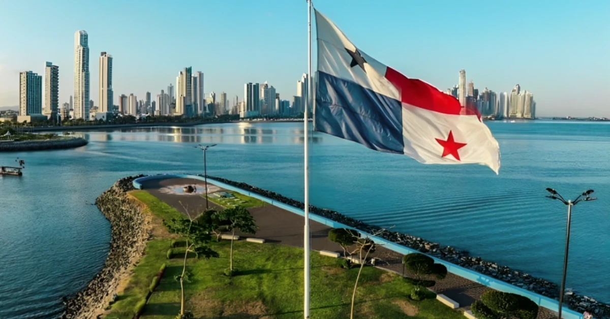 Panama flag waving above a waterfront park with a city skyline across the water.
