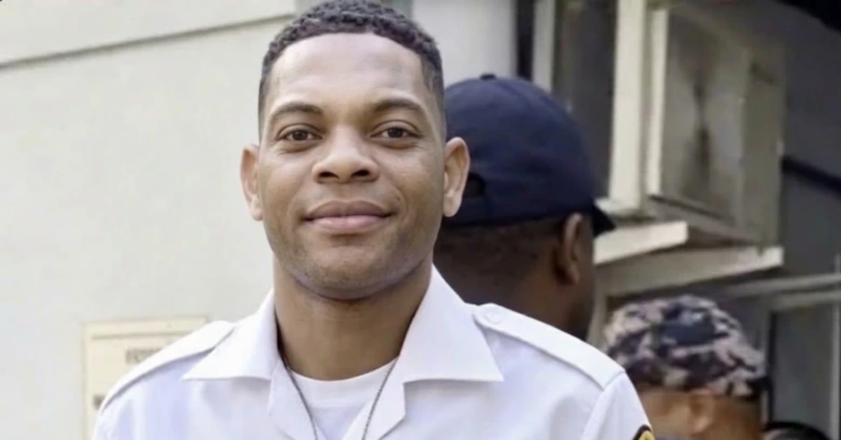 Smiling man in a light uniform outside a building, with another person wearing a cap in the background.
