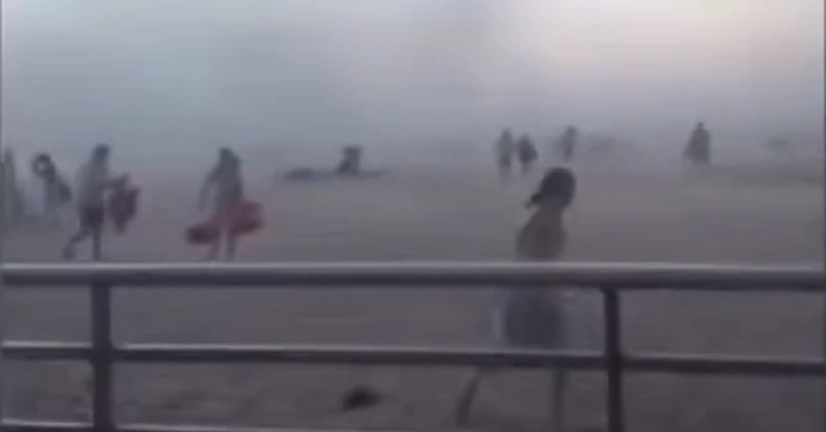 Silhouettes of people with surfboards on a foggy beach behind a railing, low visibility.