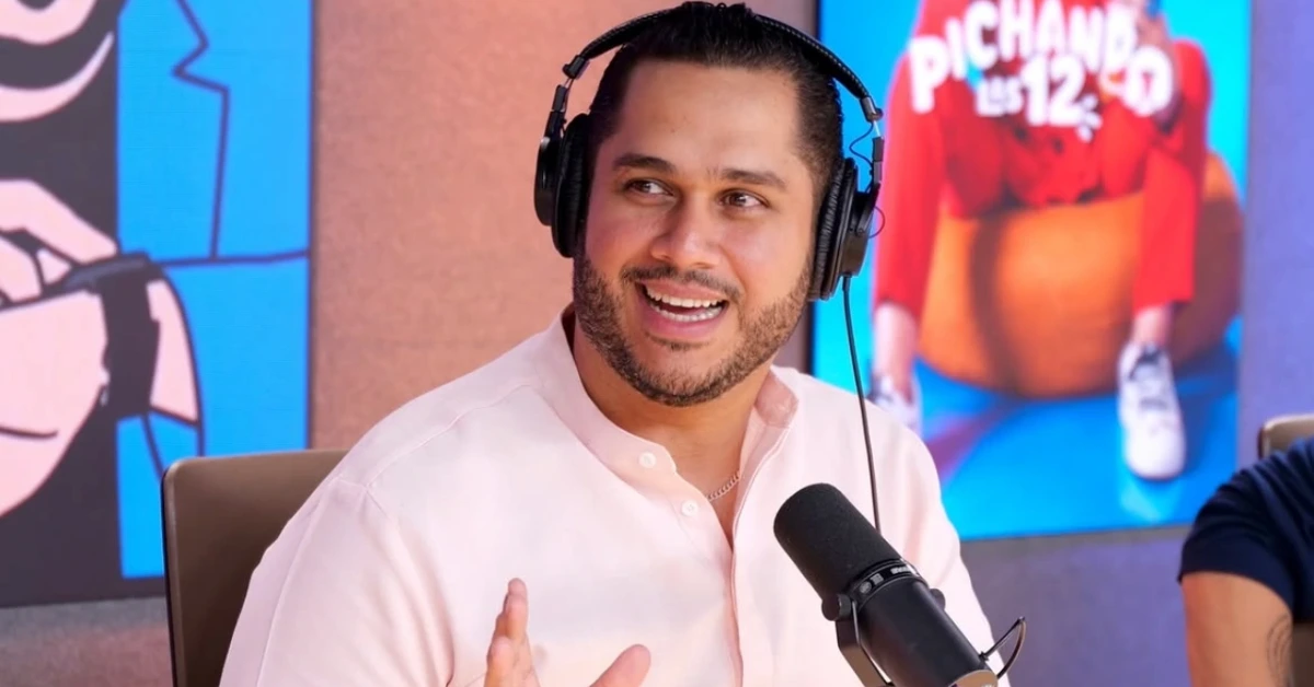 Podcast host wearing headphones, speaking into a microphone in a studio backdrop with colorful art behind him.