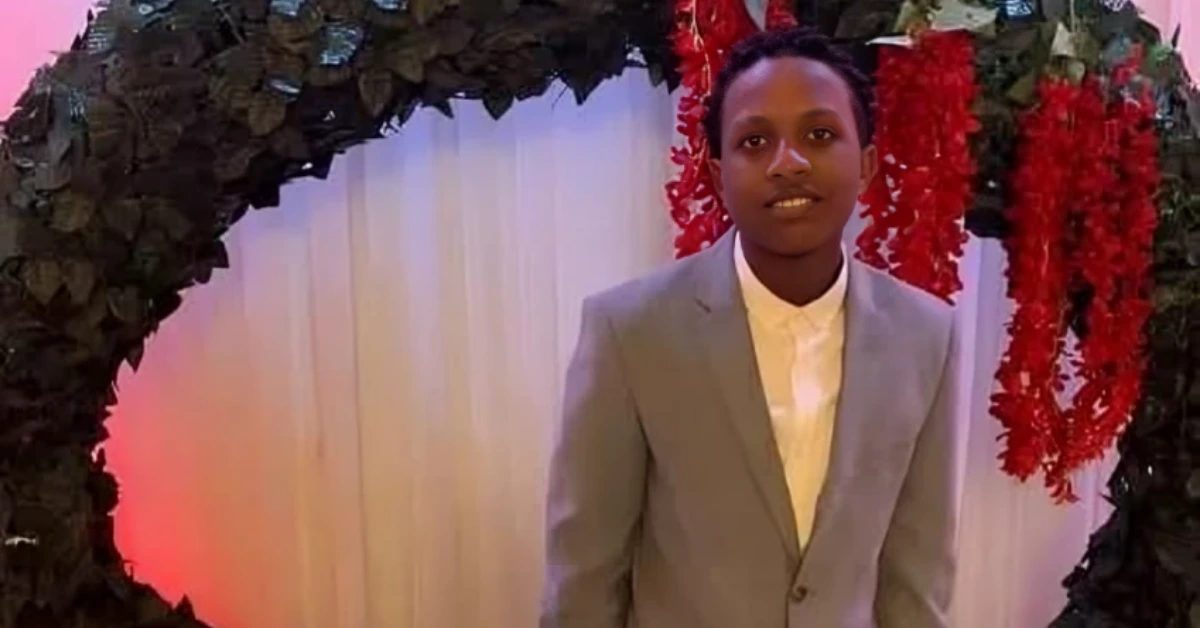 Young man in a light gray suit and white shirt stands under a leafy arch with red garlands at a formal event, posing for a photo.