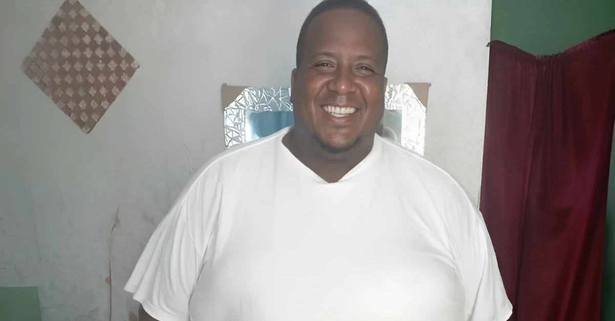 Smiling man in a white T-shirt standing indoors, with a mirror and red curtain in the background.