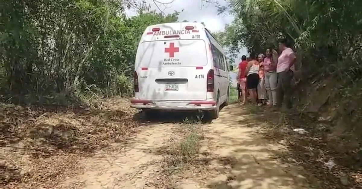 White ambulance with Red Cross emblem parked on a dirt rural road, with bystanders nearby on the right.