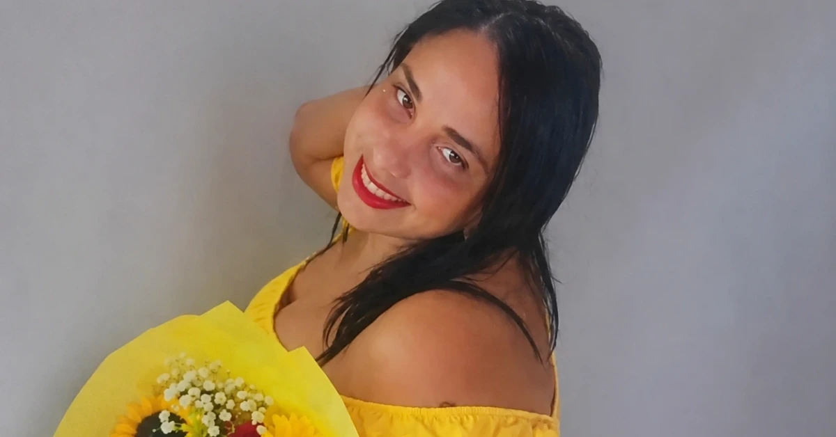 Smiling woman in a yellow off-the-shoulder dress holding a bouquet of sunflowers and baby's breath against a gray background.