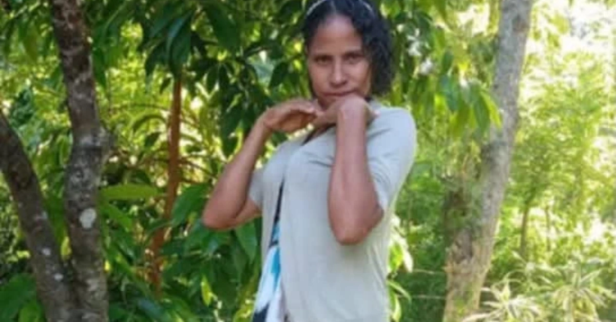 Woman posing in a leafy garden, hands near her chin, wearing a light gray top.