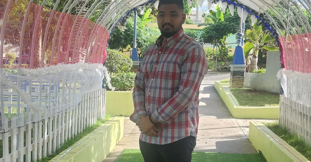 Man in a red and white plaid shirt standing with hands clasped under a decorative garden archway.