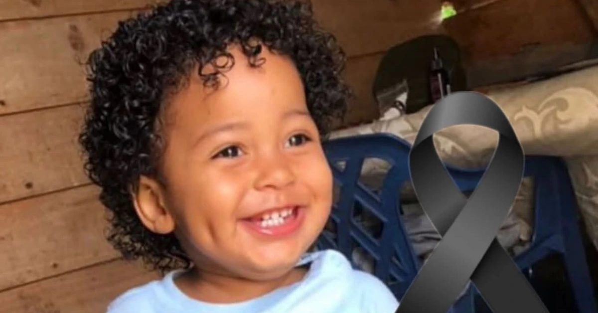 Smiling toddler with curly hair wearing a light blue shirt; a large black awareness ribbon sits beside them, signaling remembrance or support for a cause.