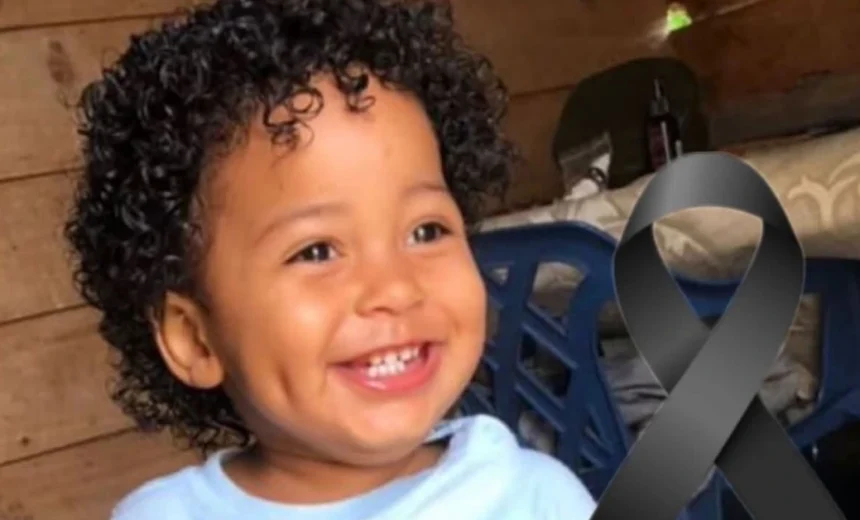 Smiling toddler with curly hair in a light blue shirt, indoors, with a large black awareness ribbon on the right side of the image.
