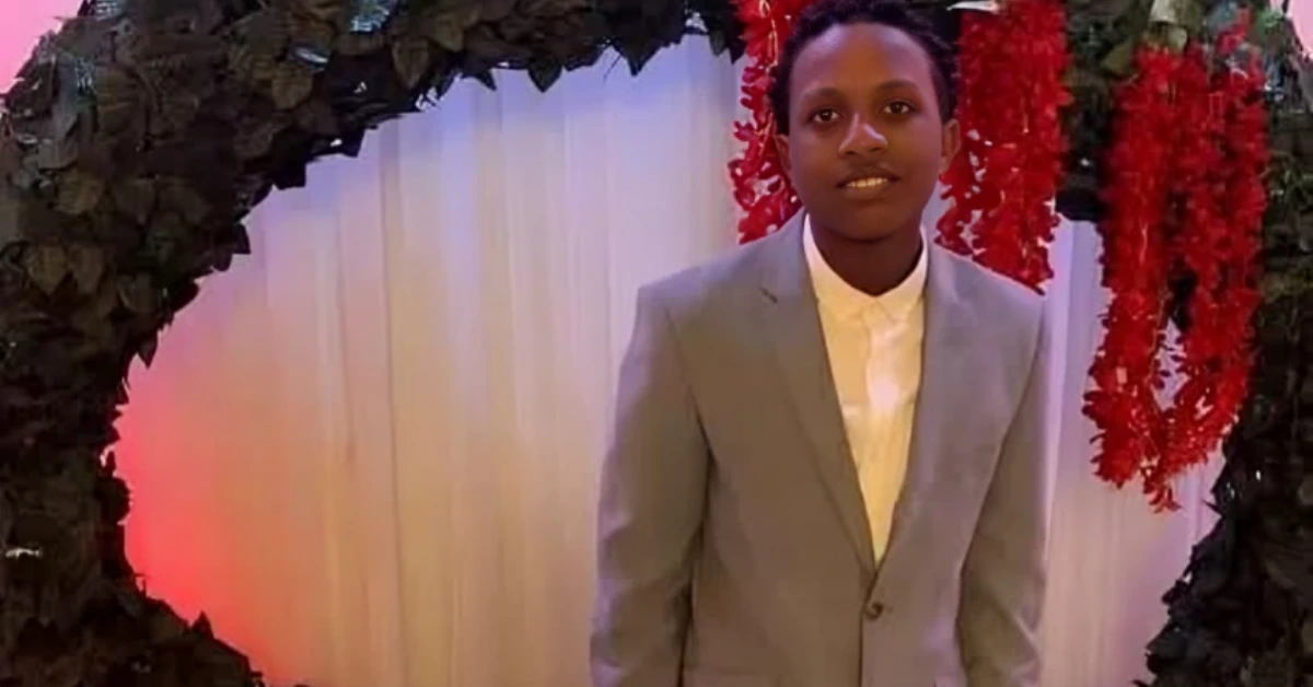 Young man in a light gray suit and white shirt posing in front of a floral arch with red garlands at an event backdrop.