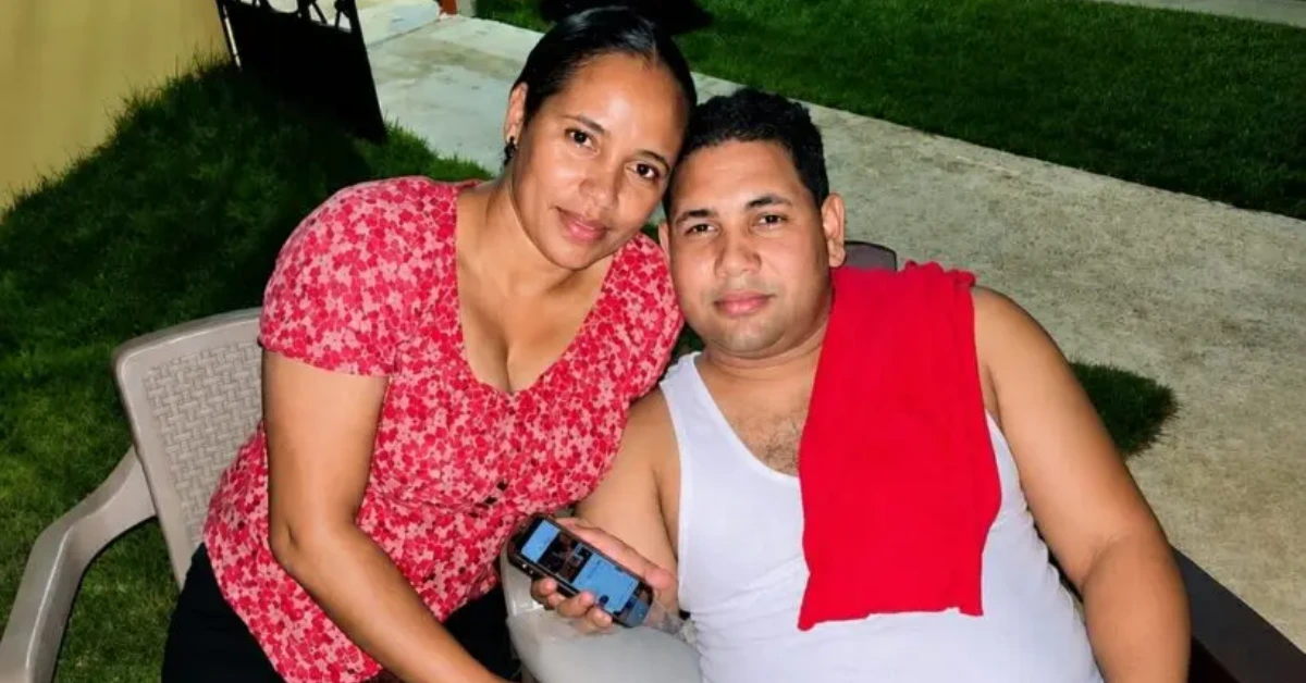 Couple sitting outdoors on lawn chairs; woman in a red floral top leaning toward man in a white tank top with a red towel over his shoulder, holding a phone.
