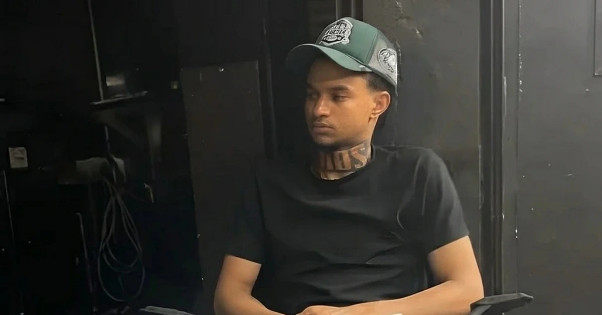 Man wearing a black T-shirt and green cap sits in a dim, backstage-like room, looking downward.
