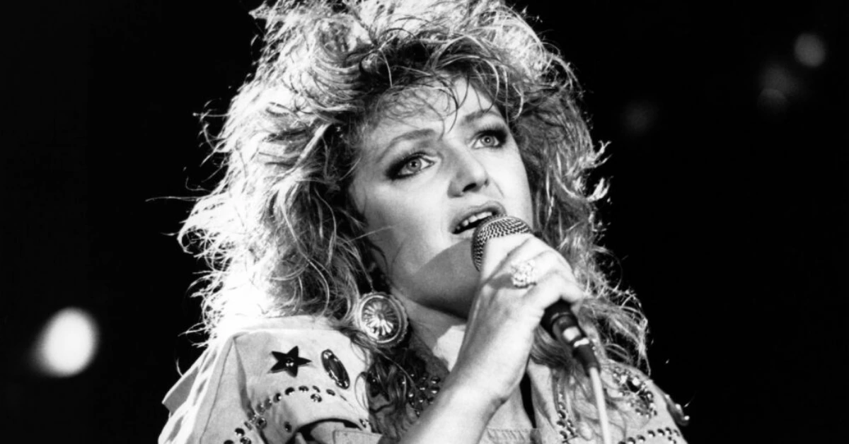 Black-and-white photo of a female singer performing into a microphone on stage, wearing a studded jacket and large earrings.