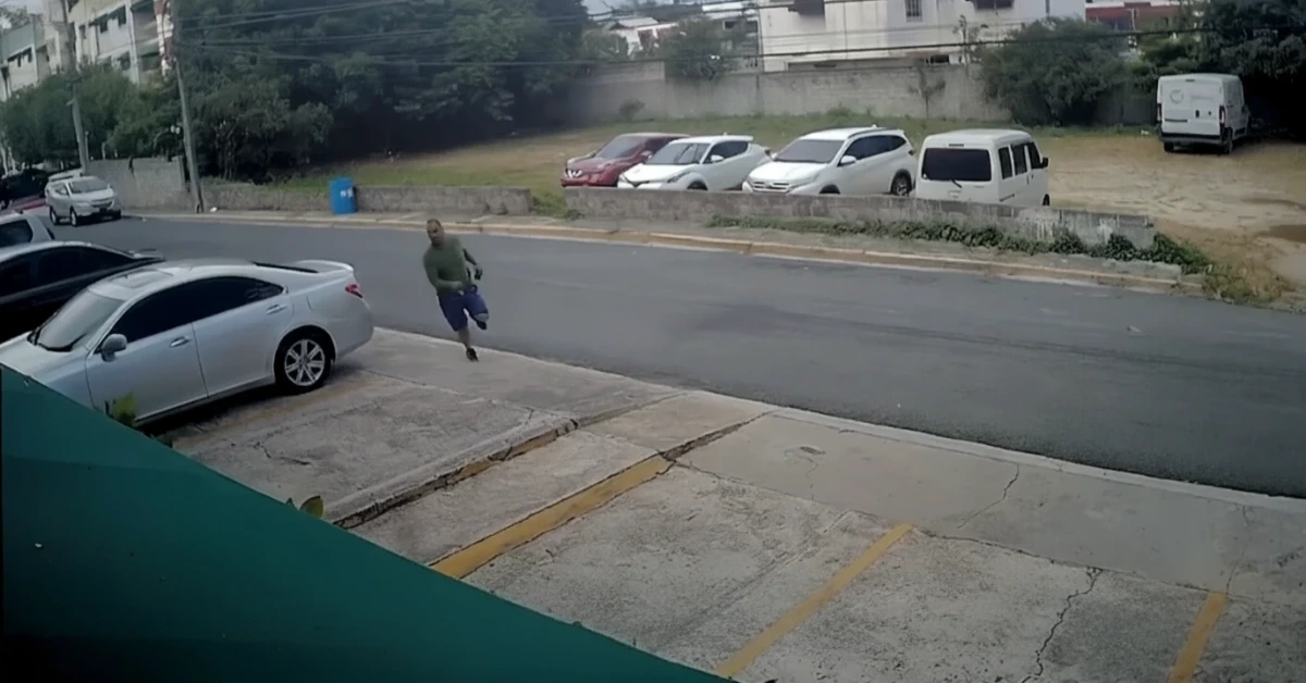 Man in a green shirt and blue shorts sprints across a sidewalk toward the street beside parked cars.