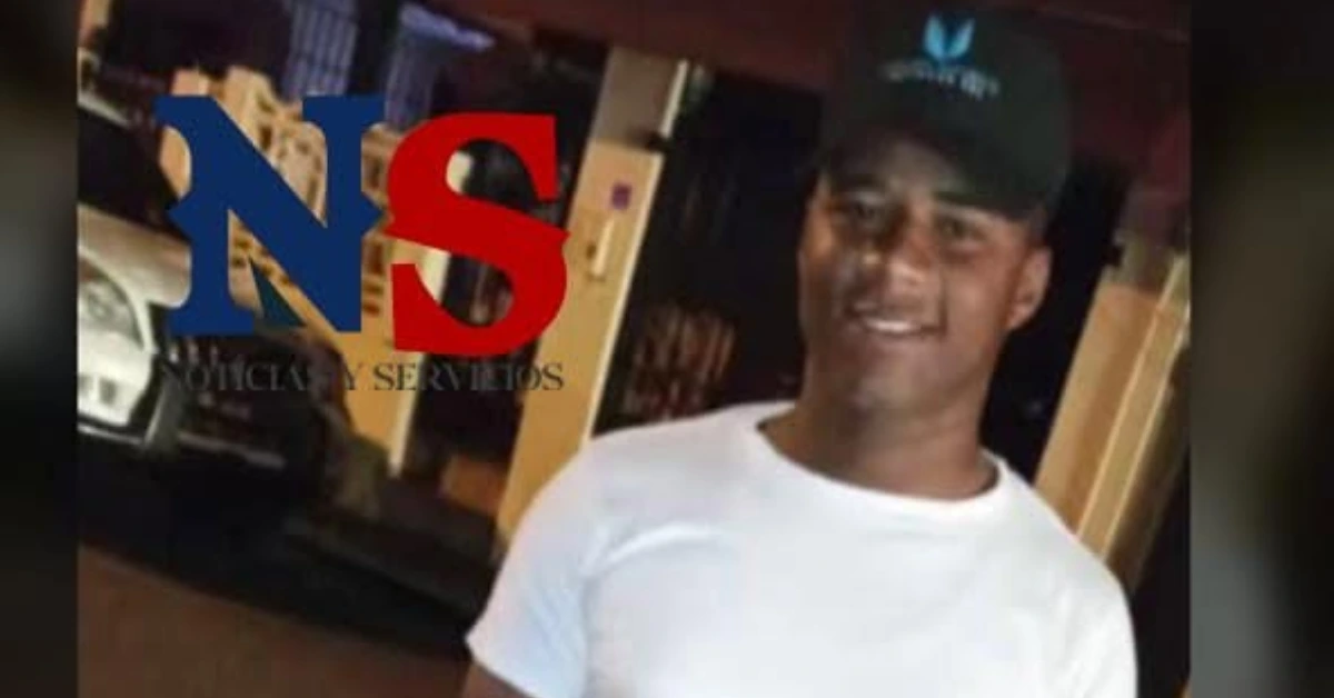 Smiling man in a white t-shirt and black cap outdoors at night, with a blue-red watermark across the image.