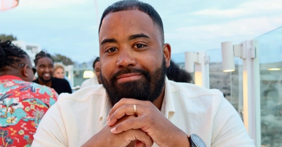 Bearded man in a light shirt sits at an outdoor dining table, hands clasped, looking at the camera.