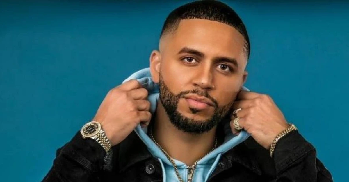 Portrait of a man with a trimmed beard wearing a blue hoodie under a black jacket, posed against a teal background in a stylish pose.