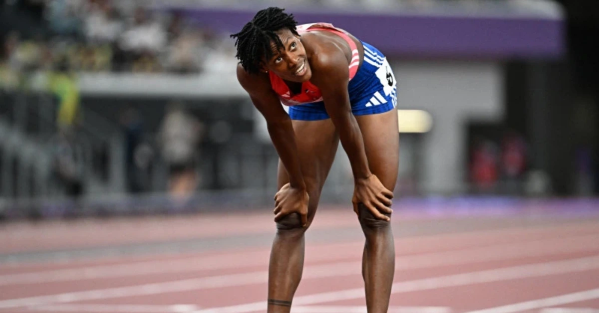 Female sprinter on a running track, hands on knees, smiling, wearing a blue and red uniform with a race bib.