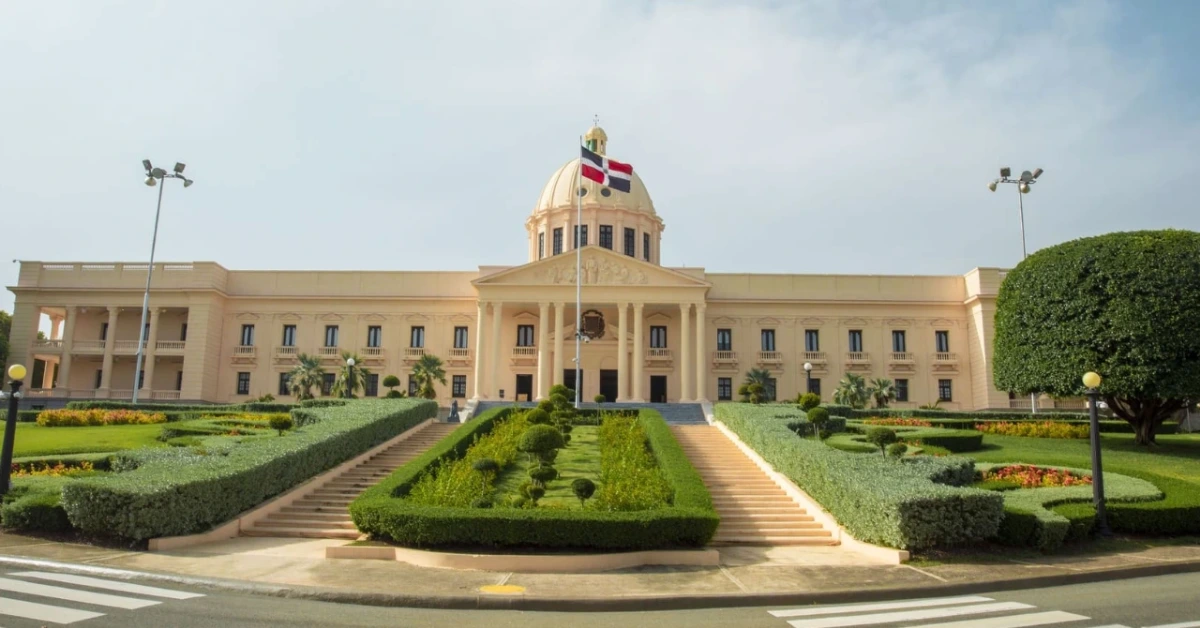 Beige neoclassical government building with a central dome, flag, and manicured garden in front.