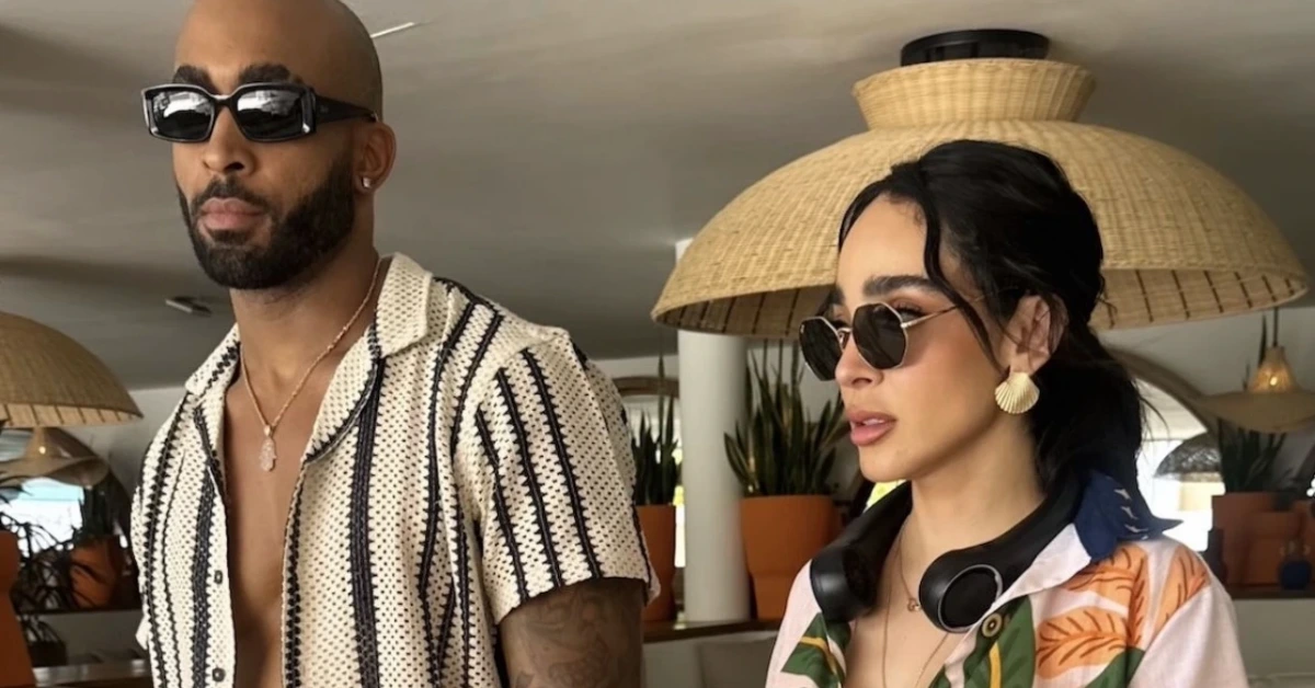 A stylish couple wearing sunglasses in a bright tropical setting, the man in a striped short-sleeve shirt and the woman in a colorful print shirt with headphones around her neck.