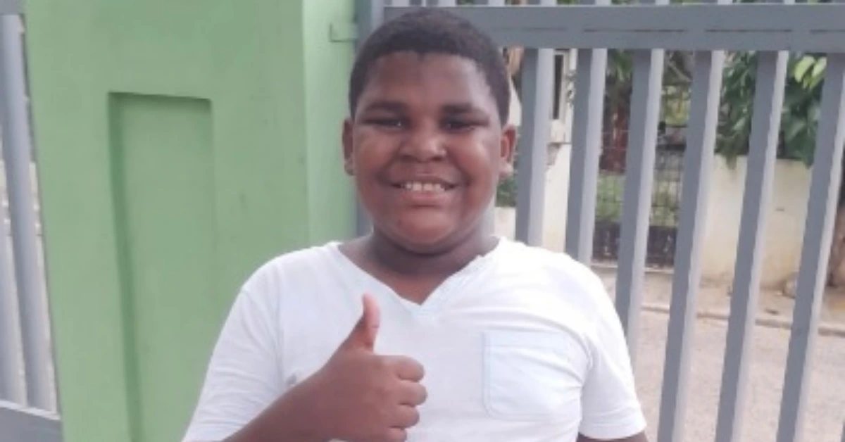 Smiling boy in a white shirt giving a thumbs up, standing outdoors beside a green wall and gray gate.