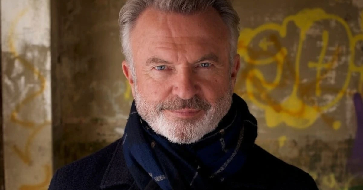Portrait of a smiling older man with gray hair and beard, wearing a dark coat and navy scarf, in front of a graffiti-covered wall.