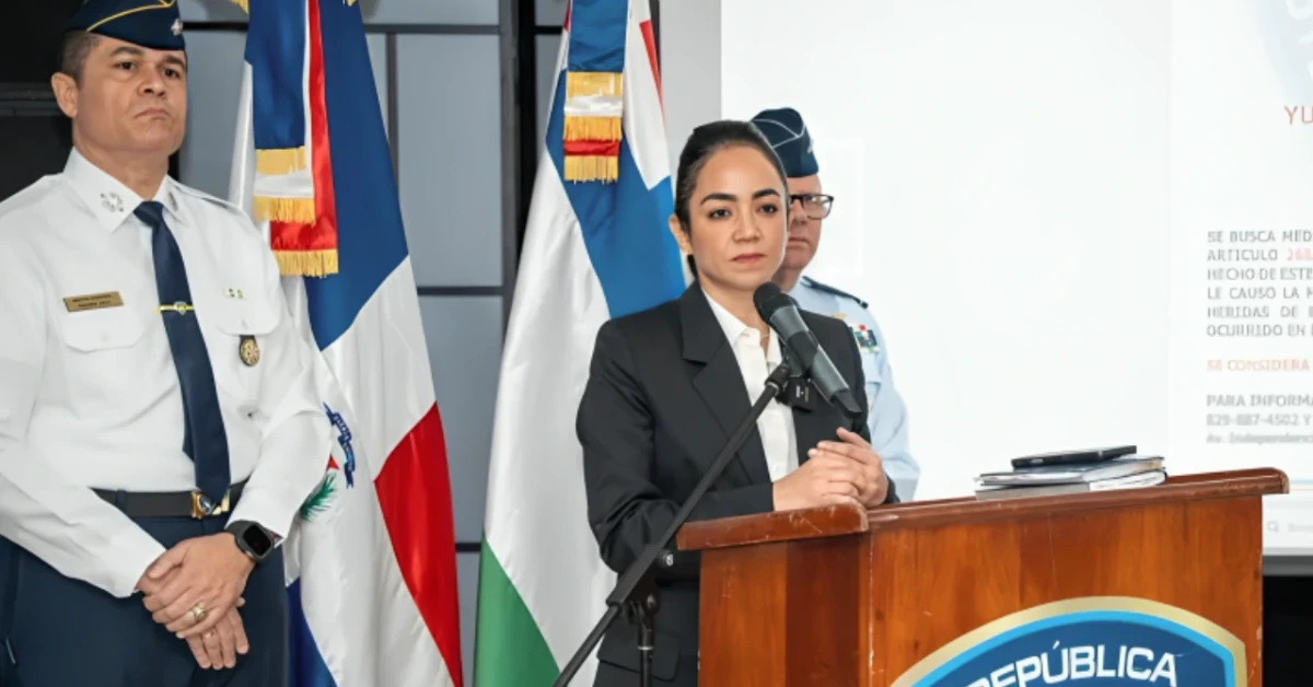 Woman in a dark suit delivers a speech at a wooden podium with a microphone; flags and uniformed officers loom in the background.