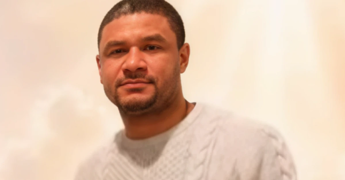 Portrait of a man with short dark hair and light stubble, wearing a gray knit sweater, looking at the camera against a soft beige background.