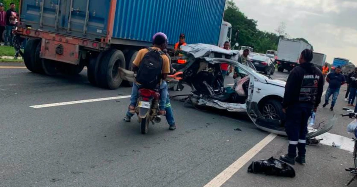 Scene of a multi-vehicle highway accident: a crushed white car, a large blue truck, and bystanders with a motorbike nearby.