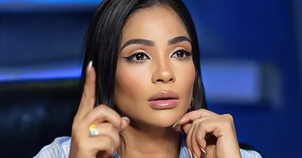 Close-up of a woman with dark hair and makeup, raising her index finger as if making a point while looking upward.