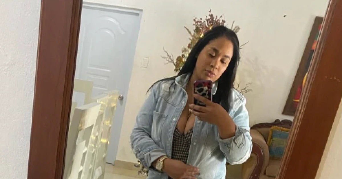 Woman taking a mirror selfie in a living room, wearing a light denim jacket over a checkered top and holding a pink leopard‑print phone.