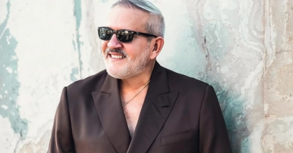Smiling man with gray hair and stubble in a dark blazer and sunglasses, leaning against a weathered wall.