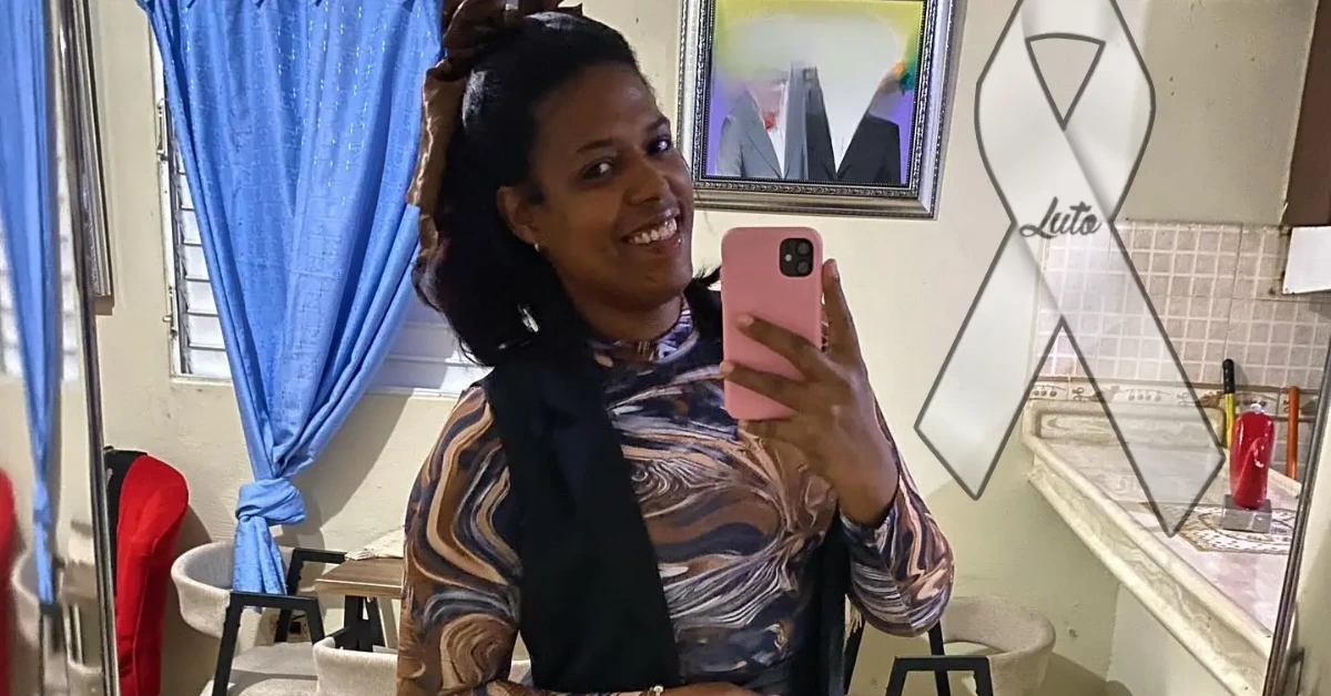 Smiling woman takes a mirror selfie in a room with a blue curtain, wearing a swirl-pattern top and black vest, holding a pink phone.