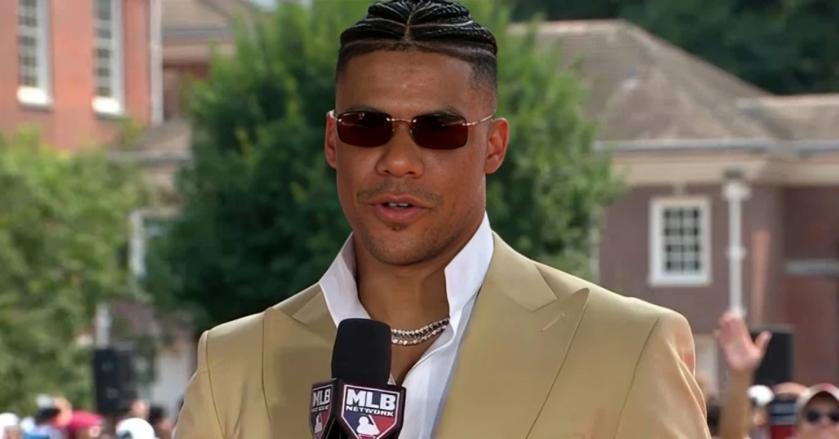 Man in a beige suit and sunglasses speaks into a branded MLB microphone at an outdoor event.