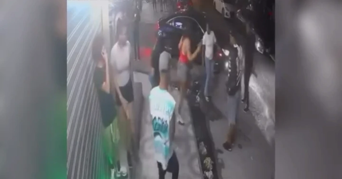 Group of people on a city sidewalk around a parked car at night, with a man in a turquoise shirt facing away from the camera in the foreground.