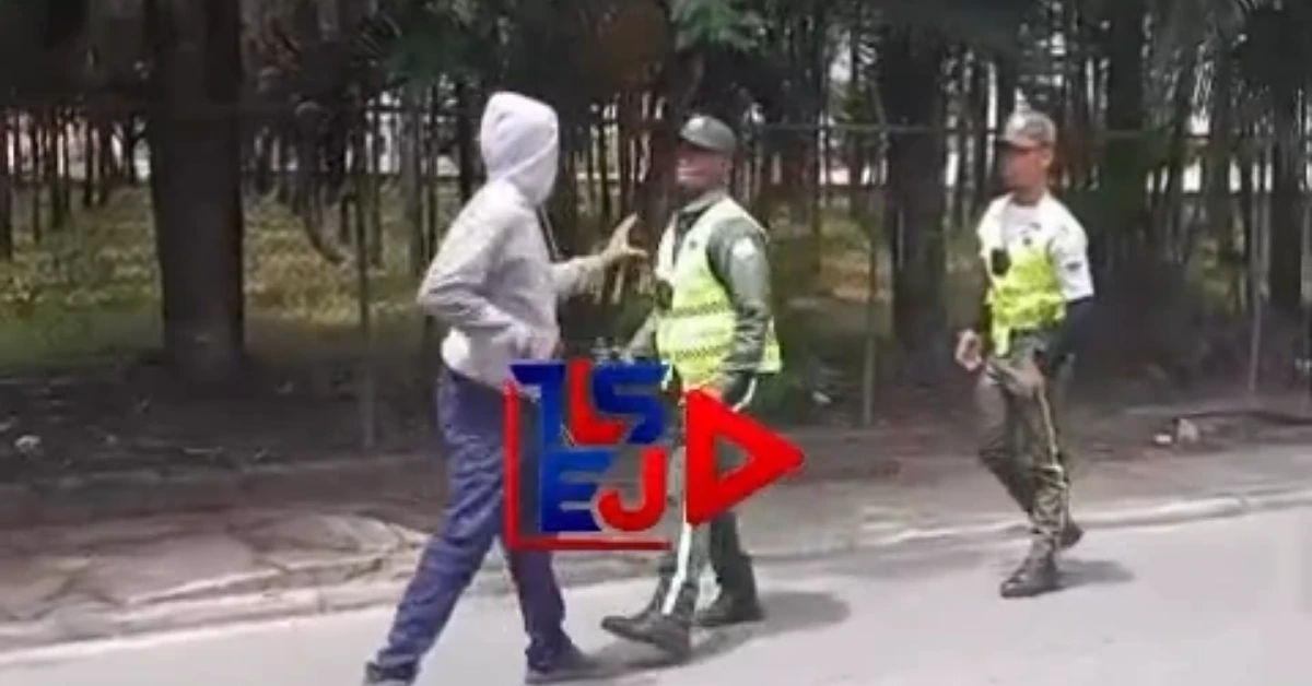 Person in a gray hooded sweatshirt talks to two police officers in high-visibility vests on a sidewalk beside a wooded area, with a colorful logo overlay in the foreground.