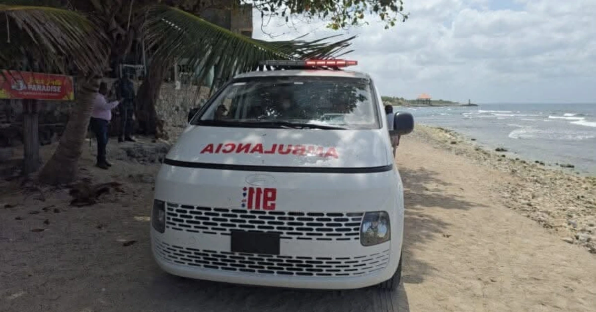 Front view of a white ambulance parked on a sandy beach with palm trees and the sea to the right, emergency lights on top.