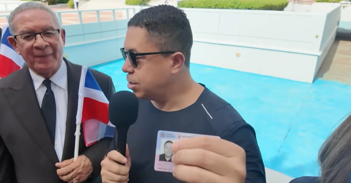 Man in a black shirt with sunglasses speaks into a handheld microphone during an outdoor interview by a blue pool, a suited man holding a flag nearby in the background.