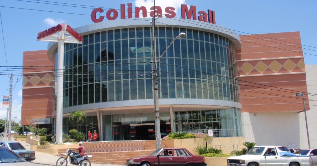 Front view of Colinas Mall, a curved glass storefront with a large red Colinas Mall sign on top and cars parked along the street.