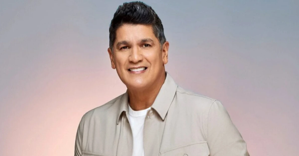 Smiling man in a beige shirt posing for a portrait against a soft pink–blue gradient background.