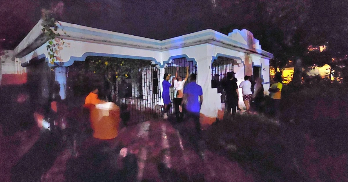 Night scene with a small white building that has blue trim, and a group of people gathered near the doorway behind barred windows.