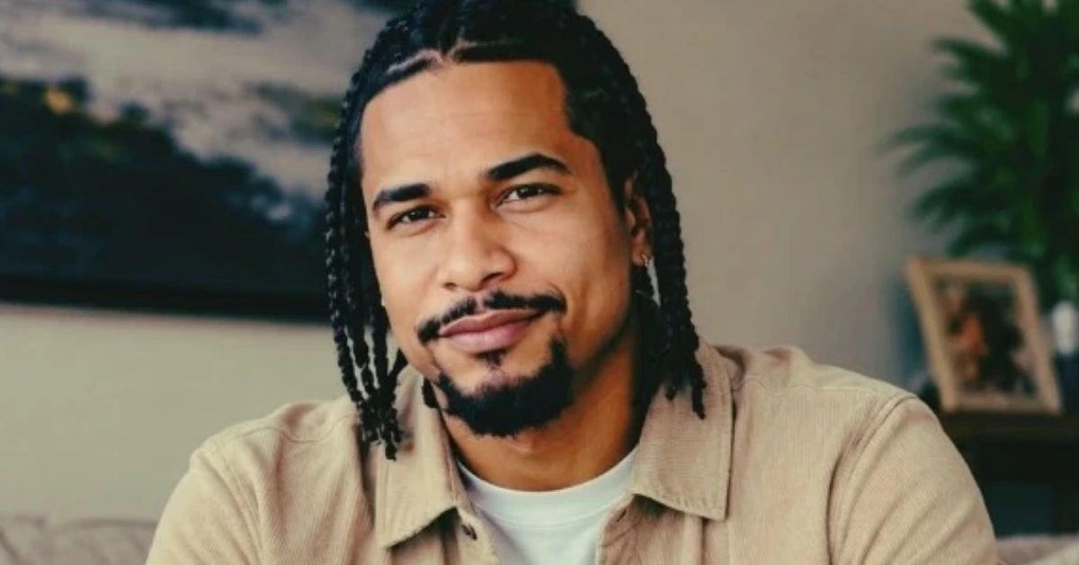 Portrait of a man with braided hair and a goatee, wearing a beige shirt and white tee, looking at the camera with a slight smile in a cozy indoor setting.